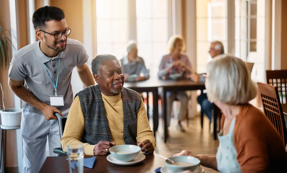 Older adult man smiling as a caregiver assists him in a dining area of an assisted living facility.