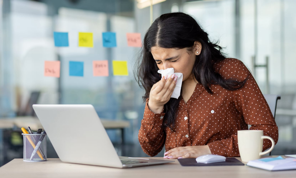 Woman blowing nose at office desk from allergies