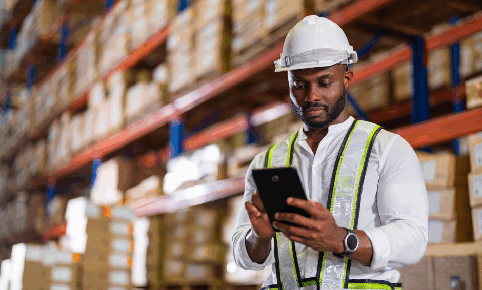 Facility manager man in warehouse with shelves of boxes using tablet with hardhat on