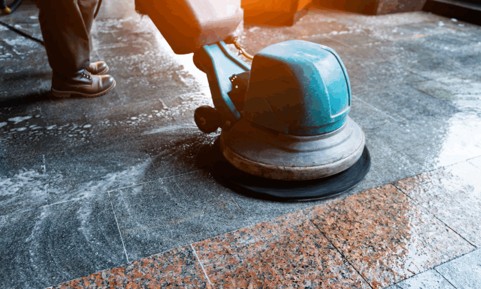 Person using a floor scrubbing machine to clean a tiled surface, leaving behind a polished shine. The image shows soap and water on the floor as the equipment works to remove dirt and grime.